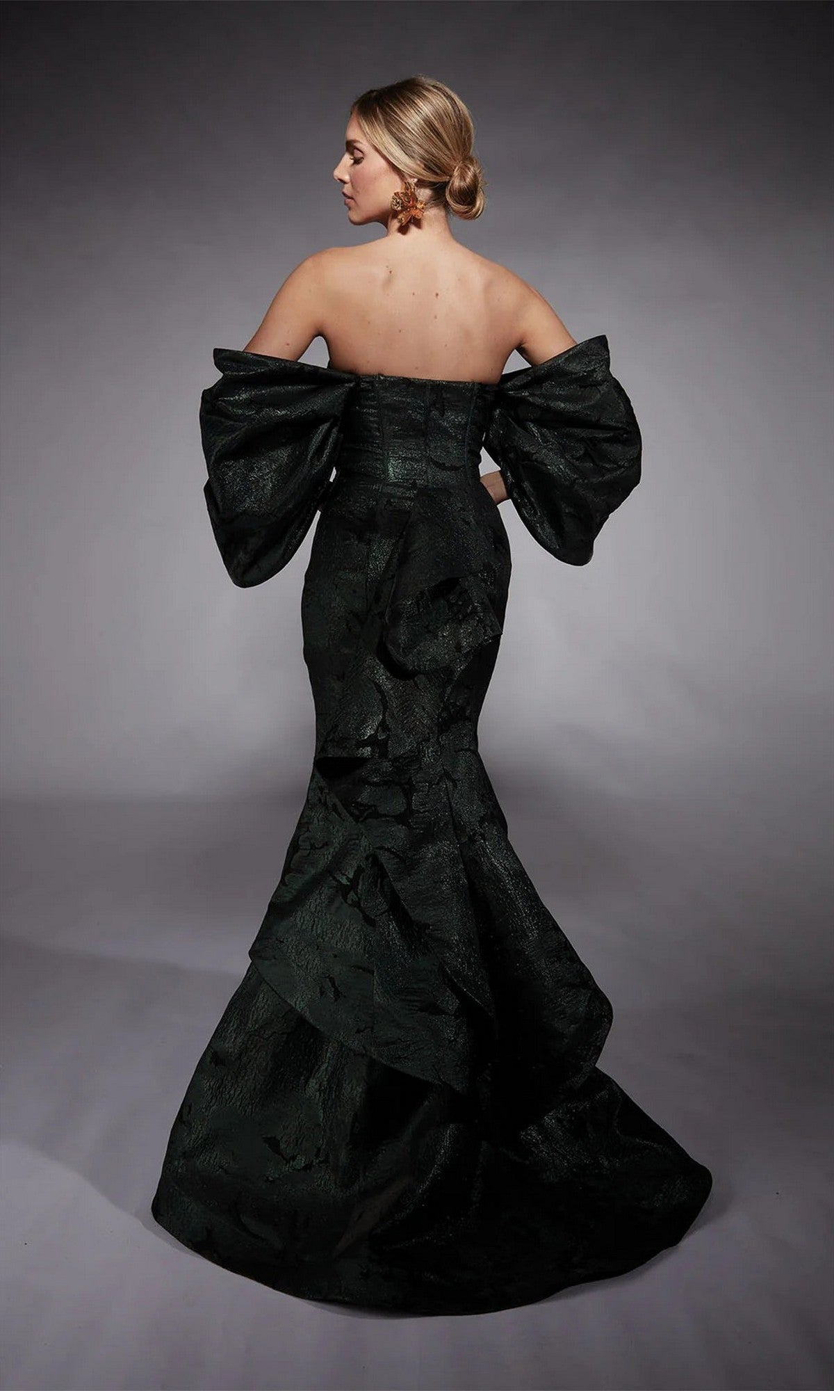 Wearing the Alyce 27747, a strapless forest green metallic jacquard corset dress with voluminous sleeves and a dramatic mermaid skirt, a woman stands in profile against a gray background.